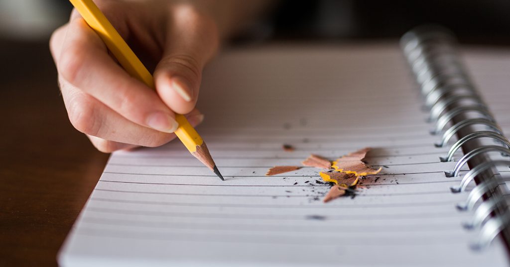Een hand die met een potlood op een schrijfblok schrijft. Er zijn ook wat schaafsels te zien en puntenslijpervulsel.