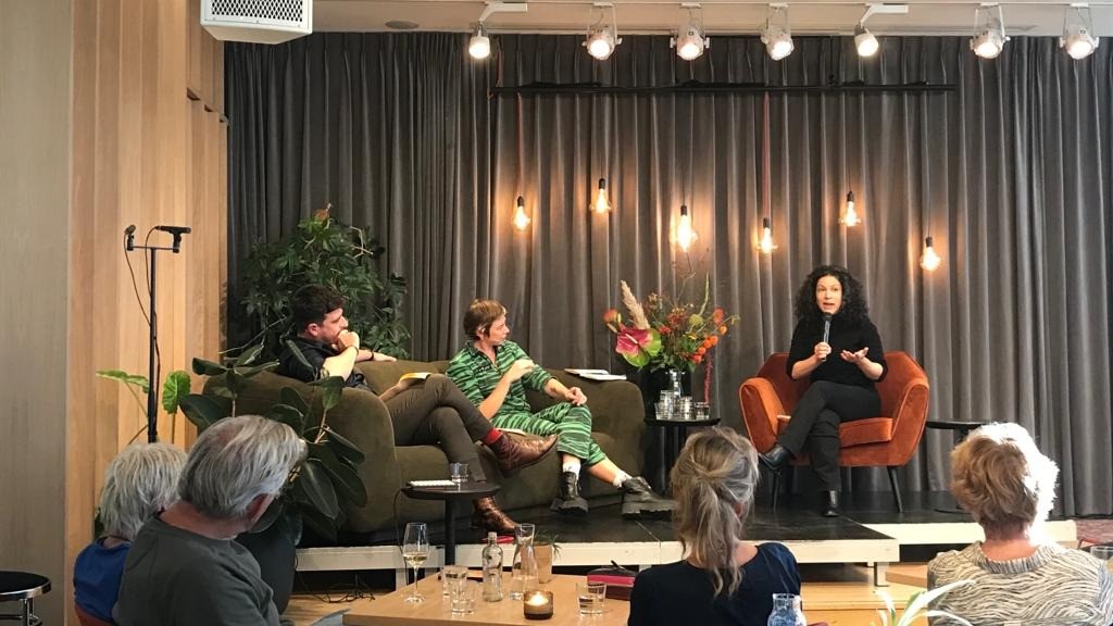 Foto van drie personen op een podium die met elkaar in gesprek zijn. Geheel rechts zit Cyrstal Hassell, coach en theatermaker. Links van haar zitten een man en een vrouw op een donkeergroene bank. Crystal spreekt in een microfoon. Achter hen hangt een grijs doen met daarvoor enkele lampen.