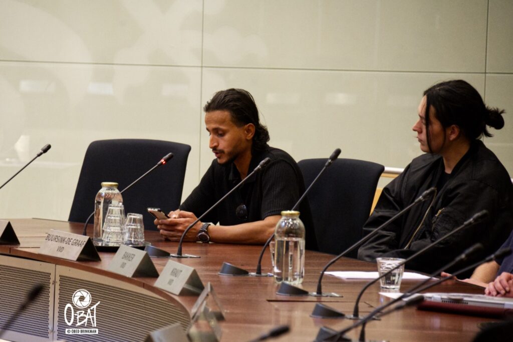 Foto van Mousa, een vluchteling uit het AZC Nijmegen Lent en een begeleidster van MiGreat die aan de inspreektafel zitten van de gemeenteraad Nijmegen in de Raadzaal in het gemeentehuis. Mousa leest voor vanaf zijn mobiel.